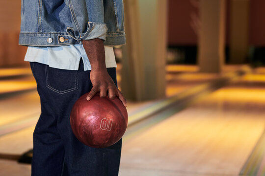 Young adult man holding bowling ball beside lane, preparing throw during leisure game. Image supporting recreation marketing, team bonding promotion, social activity campaigns