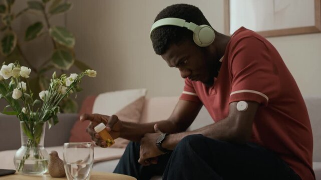 Medium full shot of young adult Black man in headphones hesitating before taking diabetes medication while sitting on couch in living room during daily care routine