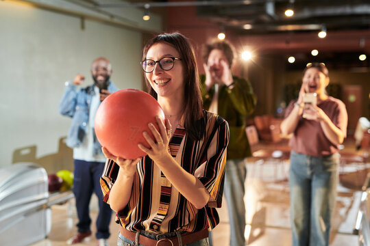 Young adult Caucasian woman holding bowling ball and preparing throw while coworkers cheering during team outing, representing workplace bonding. Ideal for team building or company culture marketing