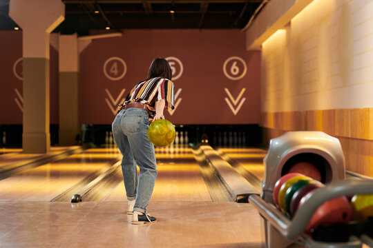 Young adult woman bowling on lane in entertainment venue, holding ball and aiming throw for leisure activity and social recreation. Useful for lifestyle, weekend fun, team bonding marketing