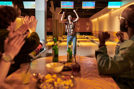 Young adult woman celebrating bowling win while friends clapping from table in alley, showing teamwork and leisure bonding. Ideal for social events, group outings, recreation marketing