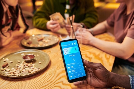 Close up of Black young adult hand holding smartphone, showing payment app while friends dining and splitting bill. Ideal for fintech, mobile banking, cashless payment marketing
