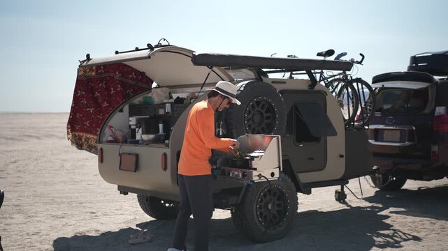 A mature man prepares a meal on a stove while camping at the beach with a teardrop camper trailer in Baja California Mexico.