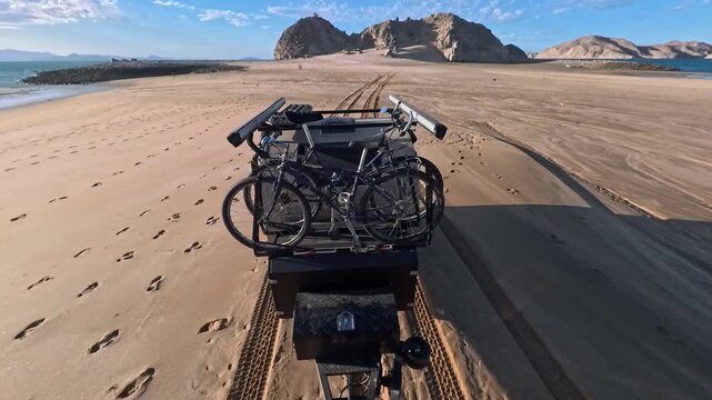 A teardrop camper travels through sand along the beach in Gonzaga Bay, Baja California, with bikes secured on the trailer.