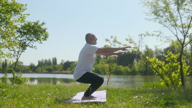 Senior man doing fitness squats exercise standing on mat in park lake