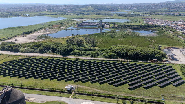 Vista a&eacute;rea de pain&eacute;is solares pr&oacute;ximos a &aacute;rea de tratamento de &aacute;gua, lago e vegeta&ccedil;&atilde;o, representando energia renov&aacute;vel e sustentabilidade ambiental.