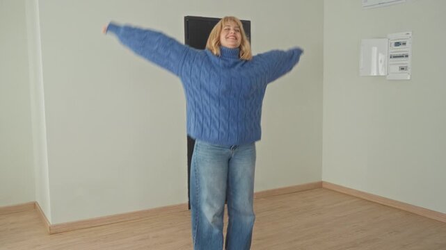 Woman in blue sweater dancing with arm raised in an empty studio room with wooden floor and visible breaker panel, wearing jeans and smiling; joyful freedom.