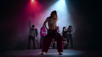 Fototapeta premium Female street dancer posing under blue spotlight with dance crew in background. Concept of dance school ad, performer casting, youth training promo and urban event visuals.