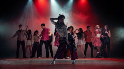 Fototapeta premium Young female dancer posing in smoky concert lights with group dancing behind. Concept of dance class campaign, stage show poster, music video casting and youth culture promo.