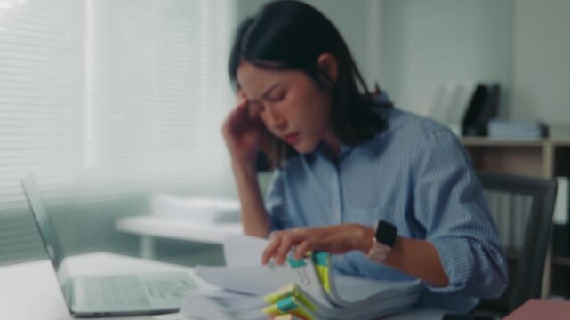 Young asian business woman feeling stressed, tired and overworked while checking a large stack of documents and reports at her office desk