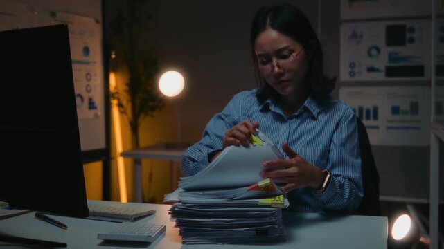 Stressed asian businesswoman working late at night in the office, meticulously sorting through a large stack of paperwork and documents