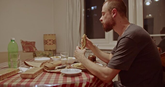 A bearded young man savors a delicious pancake, enjoying a quiet and comfortable meal alone at his dining table. Perfect for illustrating domestic life and casual evening routines.