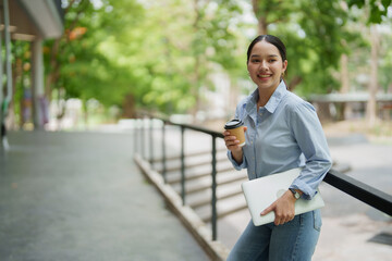 Fototapeta premium Asian university student or young freelancer smiling with a positive expression, holding a laptop and a takeaway coffee cup outdoors on a university campus or modern workspace area