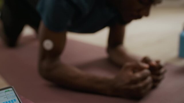 Tilt up shot of young adult Black man holding forearm plank on mat in living room while using smartphone app connected to continuous glucose monitor for diabetes care, slow motion