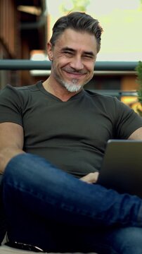Mid adult man working with phone, sitting on balcony or terrace outdoor, checking e-mail, reading news feed, posting on social media. Relaxed, confident happy smile. Businessman in home office.