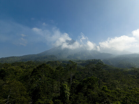 Aerial view of Mount Merapi volcano peak shrouded in clouds above a lush tropical forest under a clear blue sky in Kecamatan Cangkringan, Daerah Istimewa Yogya, Indonesia.
