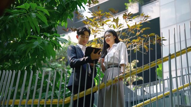 Young Asian businessman and businesswoman discussing work and looking at digital tablet while standing on outdoor staircase in modern office environment. Team collaboration and technology concept.
