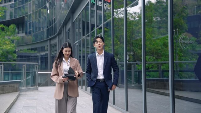Young Asian businessman and businesswoman walking and talking near modern glass office building in urban business district. Corporate lifestyle and professional teamwork concept.
