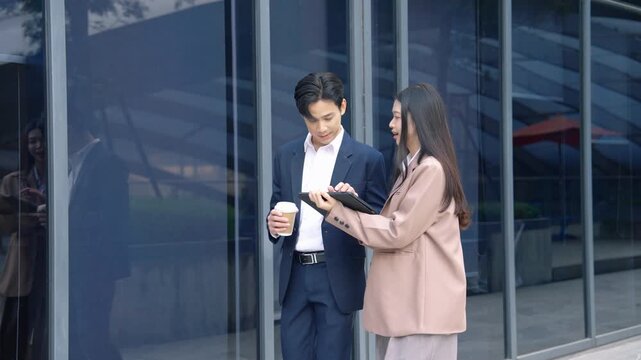 Young Asian business professionals discussing digital content on tablet device while standing outdoors in modern corporate environment. Technology and teamwork concept.
