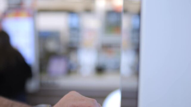 Hand taps on touch screen. Blurred background shows indoor setting. Person wears green wristband. Focus on finger movement