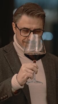 Elegant mid adult man drinking, tasting wine at home in cosy room. Portrait of happy, satisfied middle aged man, smiling.