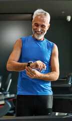 Fototapeta premium Portrait of a senior man exercising in a gym, mature male running using treadmill machine and checking smart watch for performance, healthy lifestyle and cardio exercise at fitness club concepts, vita