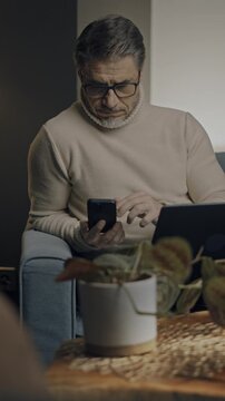 Middle aged man sitting in comfort in living room working on laptop computer and phone in home office. Businessman managing business online. Portrait of confident older male, happy, smiling.