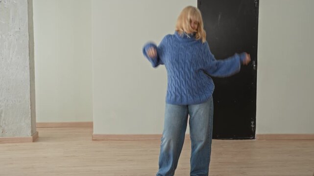 Woman in chunky blue sweater and jeans spins across an empty studio floor with arms raised and a bright smile, mid dance; joyful movement.