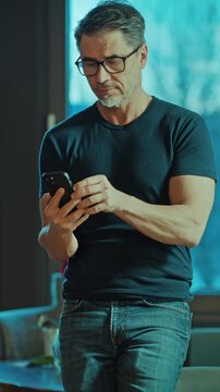 Happy, confident older man standing in living room at home using phone, smiling. Mature age, middle age, mid adult casual guy in 50s, bearded, gray hair.