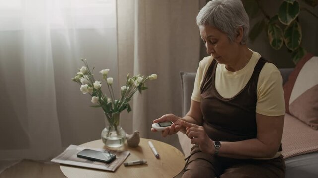 Pull back shot of mature woman performing fingerstick blood test on glucometer while managing diabetes routine sitting at coffee table in living room