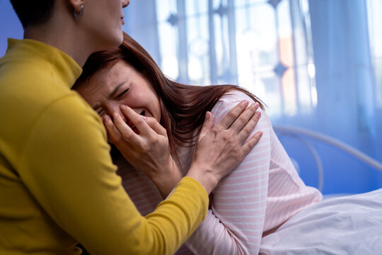 Young short haired woman comforting and supporting her crying friend on bed at home