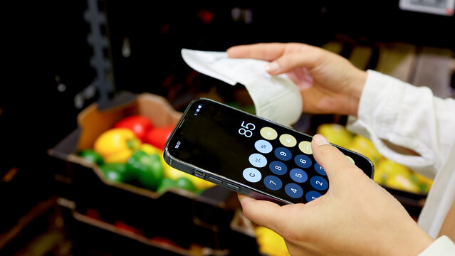 Female shopper checking receipt and using calculator in supermarket aisle with products in background. Concept expense control and consumer behavior.
