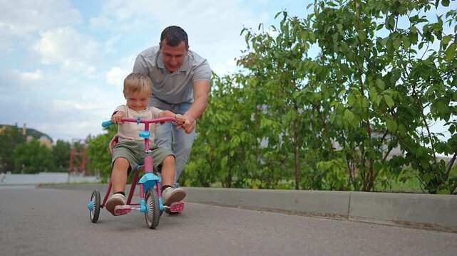 Father helps his son ride tricycle on paved path. Happy family walks with little boy on bike outdoors. Dad pushes happy child on trike in summer. Father teaching kid to ride bike enjoys sunny nature.