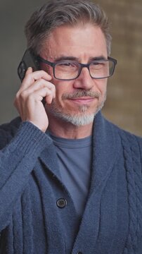 Middle aged man on phone call at home. Gray haired older male in glasses and sweater talking on mobile in living room. Telecommunication .