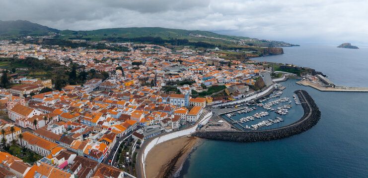Aerial view of the historic city center with orange-tiled rooftops, the harbor filled with boats, and the Se Cathedral under a cloudy sky in Angra do Heroismo, Azores, Portugal.