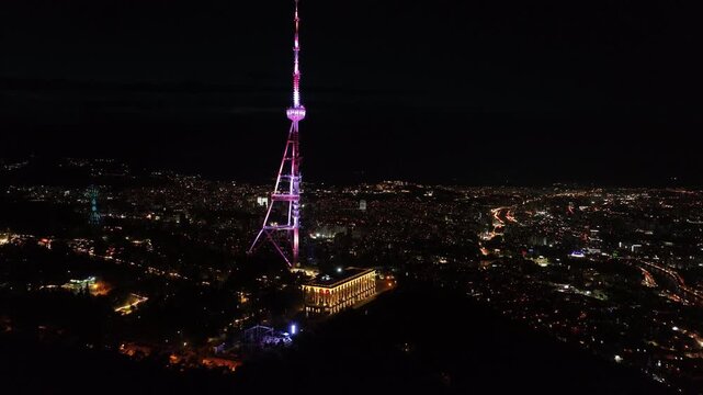 Aerial view of Tbilisi TV Tower illuminated at night with city lights and Holy Trinity Cathedral in the background, Georgia cityscape from Mtatsminda Park at dusk.