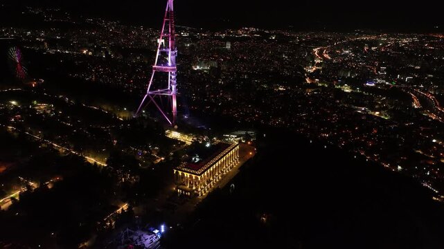 Aerial view of Tbilisi TV Tower illuminated at night with city lights and Holy Trinity Cathedral in the background, Georgia cityscape from Mtatsminda Park at dusk.