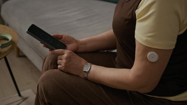 OTS shot of middle aged woman wearing continuous glucose monitor on arm asking AI assistant about diabetes question while sitting on couch in living room, dolly away camera movement