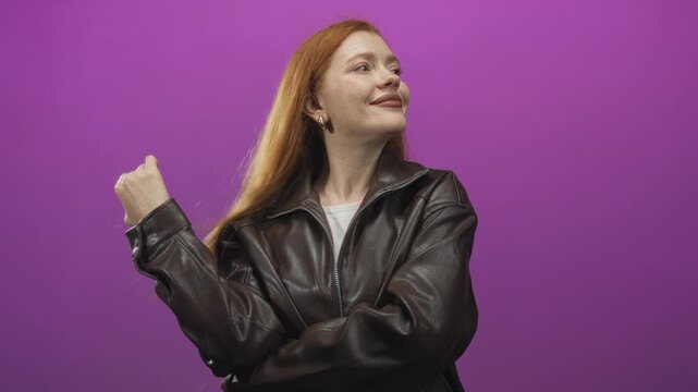 Redhead woman touches hair and smiles, wearing brown leather jacket with crossed arms and tilted head in studio against purple backdrop; confidence self care.