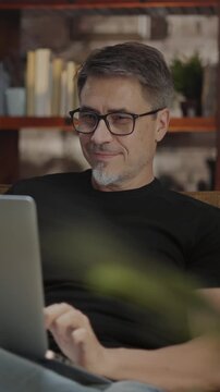 Older man working online with laptop computer at home sitting on couch in cosy living room. Home office, browsing internet. Portrait of happy, mature age, middle age, mid adult man in 50s, smiling.