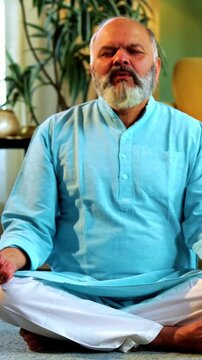 Indian senior couple doing omkar pranayama or yoga at home in the morning sitting with folded legs in modern living room chanting aum, practicing mindful breathing exercise for health