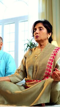 Indian senior couple doing omkar pranayama or yoga at home in the morning sitting with folded legs in modern living room chanting aum, practicing mindful breathing exercise for health