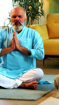 Indian senior couple doing omkar pranayama or yoga at home in the morning sitting with folded legs in modern living room chanting aum, practicing mindful breathing exercise for health