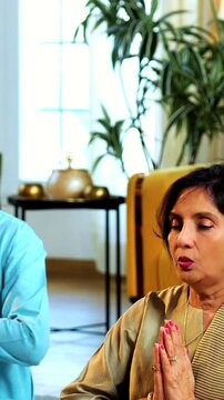 Indian senior couple doing omkar pranayama or yoga at home in the morning sitting with folded legs in modern living room chanting aum, practicing mindful breathing exercise for health