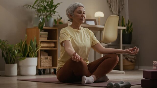 Full length shot of senior woman wearing continuous glucose monitor on upper arm sitting in lotus pose on mat while meditating during home yoga session
