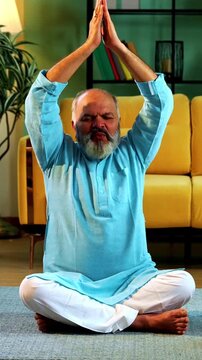 Indian senior couple doing omkar pranayama or yoga at home in the morning sitting with folded legs in modern living room chanting aum, practicing mindful breathing exercise for health