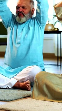 Indian senior couple doing omkar pranayama or yoga at home in the morning sitting with folded legs in modern living room chanting aum, practicing mindful breathing exercise for health
