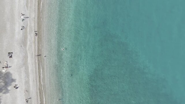 Vertical top-down drone view of the crystal-clear turquoise tide washing onto a sandy beach. The footage captures the rhythmic white foam of waves and people relaxing on the shore, highlighting the se