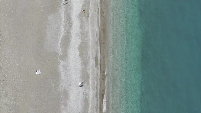 Vertical top-down drone view of the crystal-clear turquoise tide washing onto a sandy beach. The footage captures the rhythmic white foam of waves and people relaxing on the shore, highlighting the se