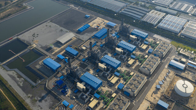 Aerial view of the Gulf Pluak Daeng Power Plant featuring industrial buildings with blue roofs, tall smokestacks, and cooling towers under clear sunlight in Map Yang Phon, Rayong, Thailand.